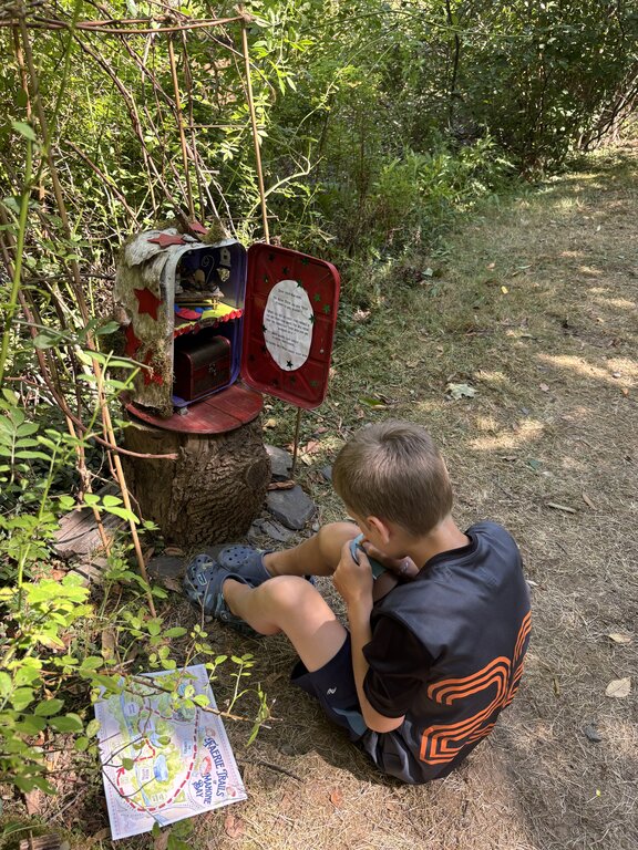 A young boy sits on a forest path examining a small item beside a whimsical fairy mailbox made from a painted red box on a tree stump. A colourful map of the faerie trail lies on the ground nearby.