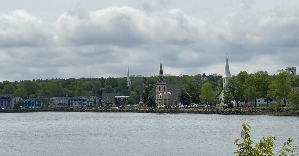 A panoramic view of Mahone Bay, Nova Scotia, shows its famous three waterfront churches with white steeples rising above the tree line under a cloudy sky.