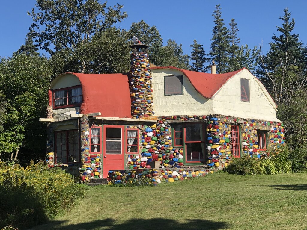 A whimsical two-story house with a red roof and multicolored stones covering its exterior walls and chimney sits in a grassy clearing surrounded by trees. The bold design and hand-painted stones give the home a storybook feel.