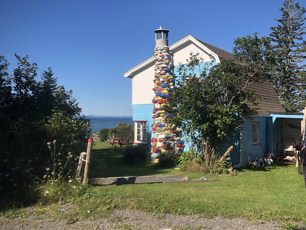 A coastal cottage with bright blue trim and a tall stone chimney painted in vibrant colors stands against a backdrop of blue sky and ocean in Nova Scotia. The playful design adds charm to the lush green lawn and seaside setting.