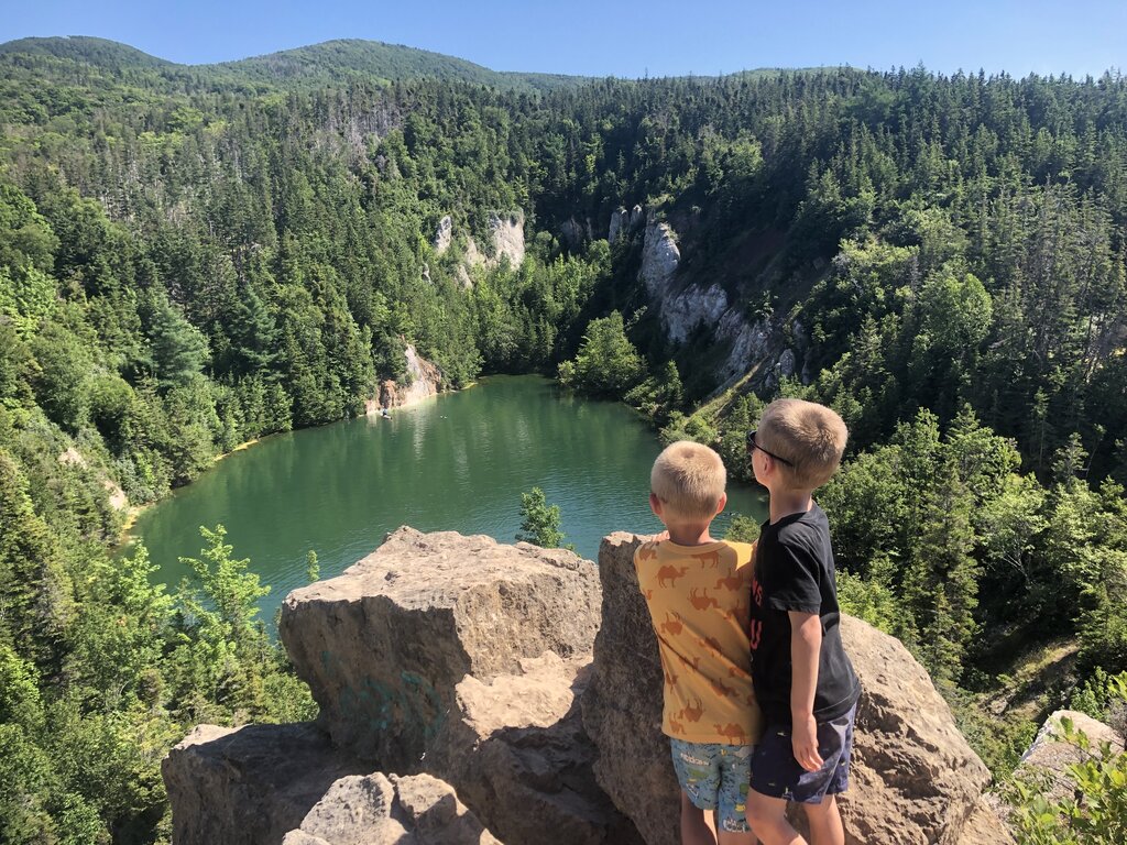Two children stand on a rocky cliff overlooking a green lake surrounded by forested hills under a clear blue sky.