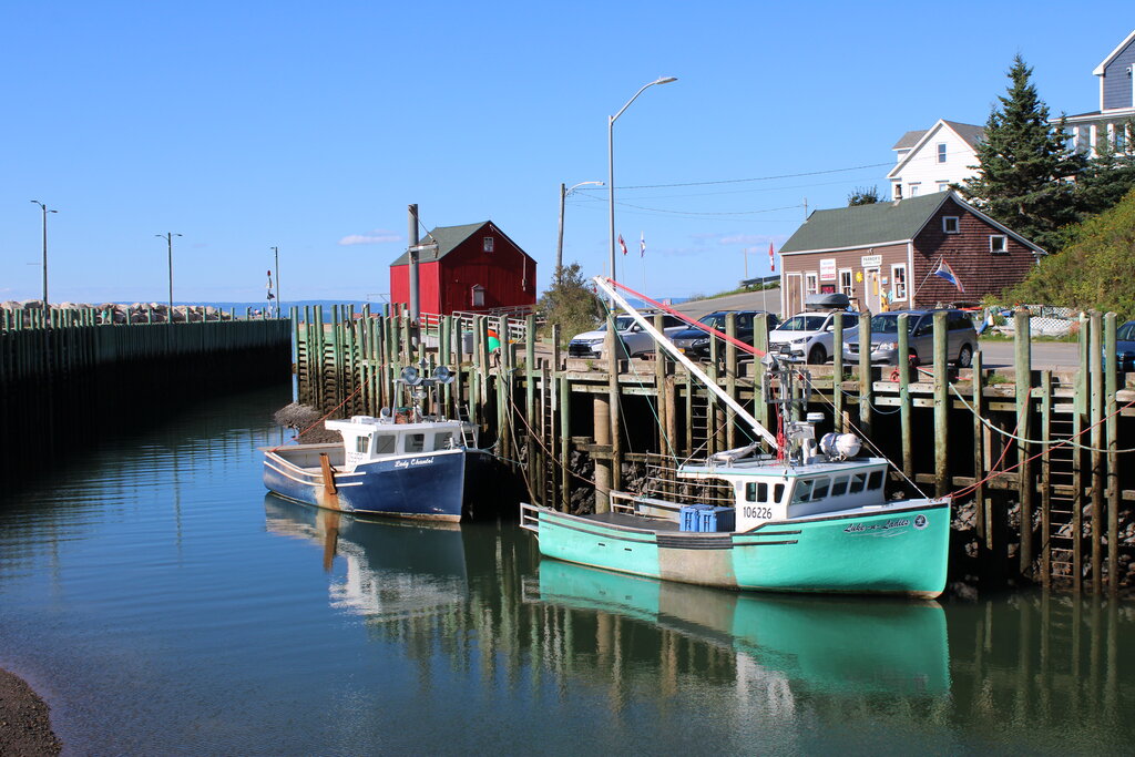 Two fishing boats sit in a quiet harbour at Hall’s Harbour in Nova Scotia with wooden docks, coastal buildings and a red structure along the shoreline.