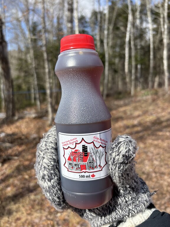 A gloved hand holds a plastic bottle of maple syrup outdoors in the woods. The label reads "Pure Nova Scotia MAPLE SYRUP" and "Sirop d érable de la Nouvelle Écosse" with "500 mL" printed at the bottom.