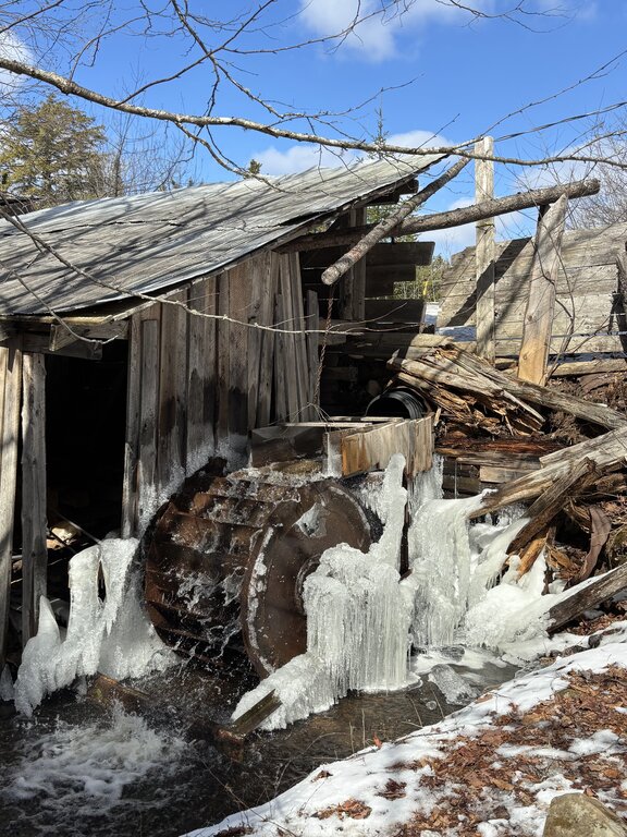 A weathered wooden structure with a water wheel sits beside a partially frozen stream with icicles hanging from the machinery. This rustic scene shows traditional maple syrup Nova Scotia production during early spring thaw.