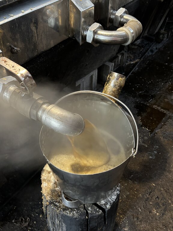 Hot maple syrup pours from a metal pipe into a bucket inside the sugar shack with steam rising around it. This close up captures the final stage of collecting maple syrup Nova Scotia as it flows from the evaporator.