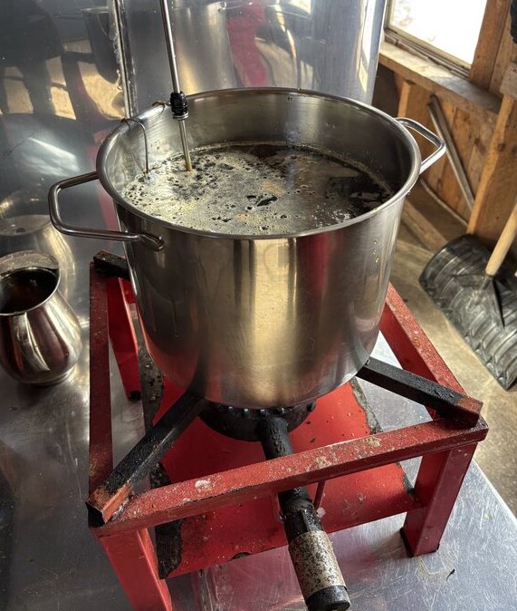 A large stainless steel pot of dark boiling syrup sits on a red gas burner inside the sugar shack. Steam rises from the surface as the maple syrup cooks down.