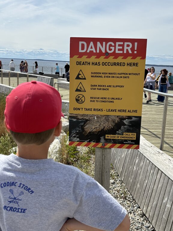 Child in a red hat looking at a yellow and red warning sign beside a coastal boardwalk. The sign reads “DANGER. Death has occurred here” and warns about sudden high waves, slippery rocks, and difficult rescue conditions.