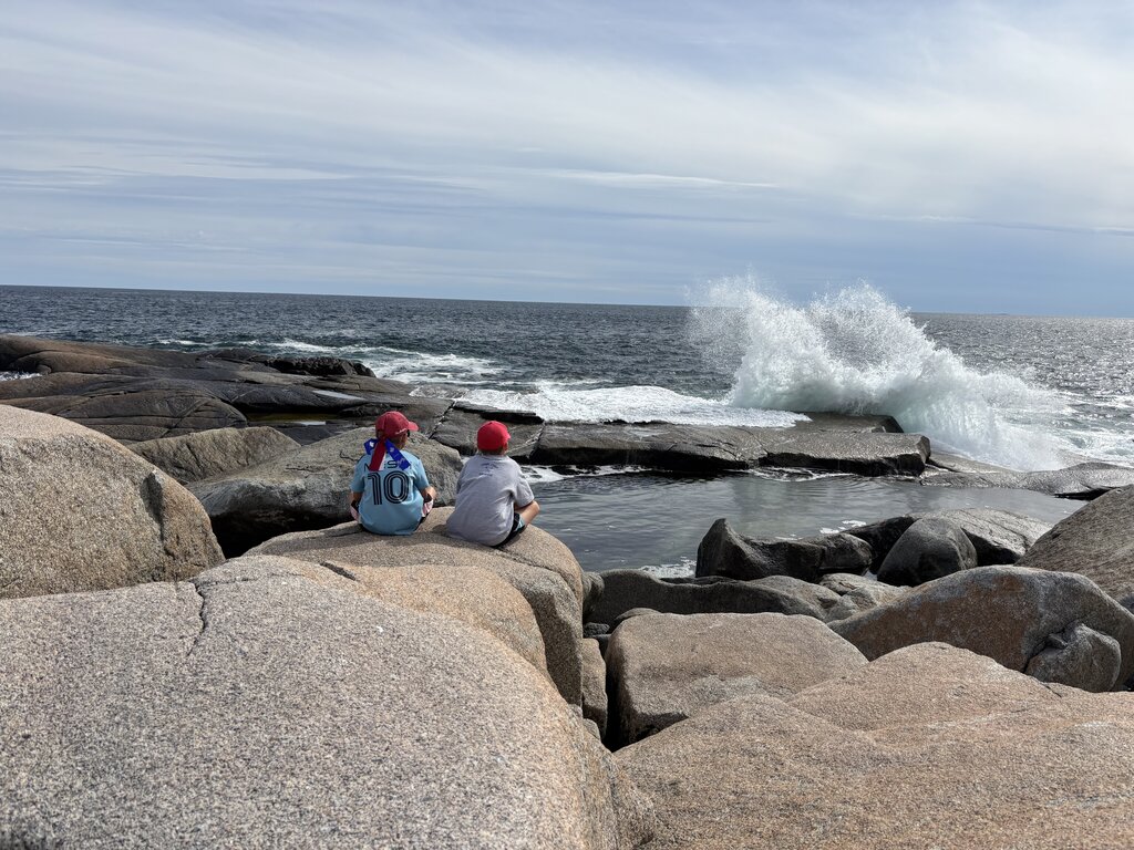 Two children sitting on large granite rocks watching ocean waves crash against the rocky shoreline at Peggy's Cove Nova Scotia. White spray rises from the water beneath a cloudy coastal sky.