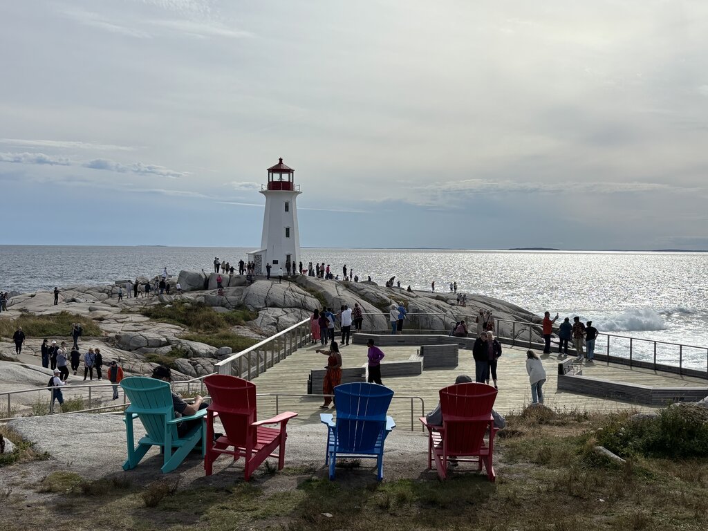 The white lighthouse of Peggy's Cove standing on rocky coastline beside a busy viewing platform filled with visitors and colourful Adirondack chairs facing the ocean. Sunlight reflects across the water behind the lighthouse.