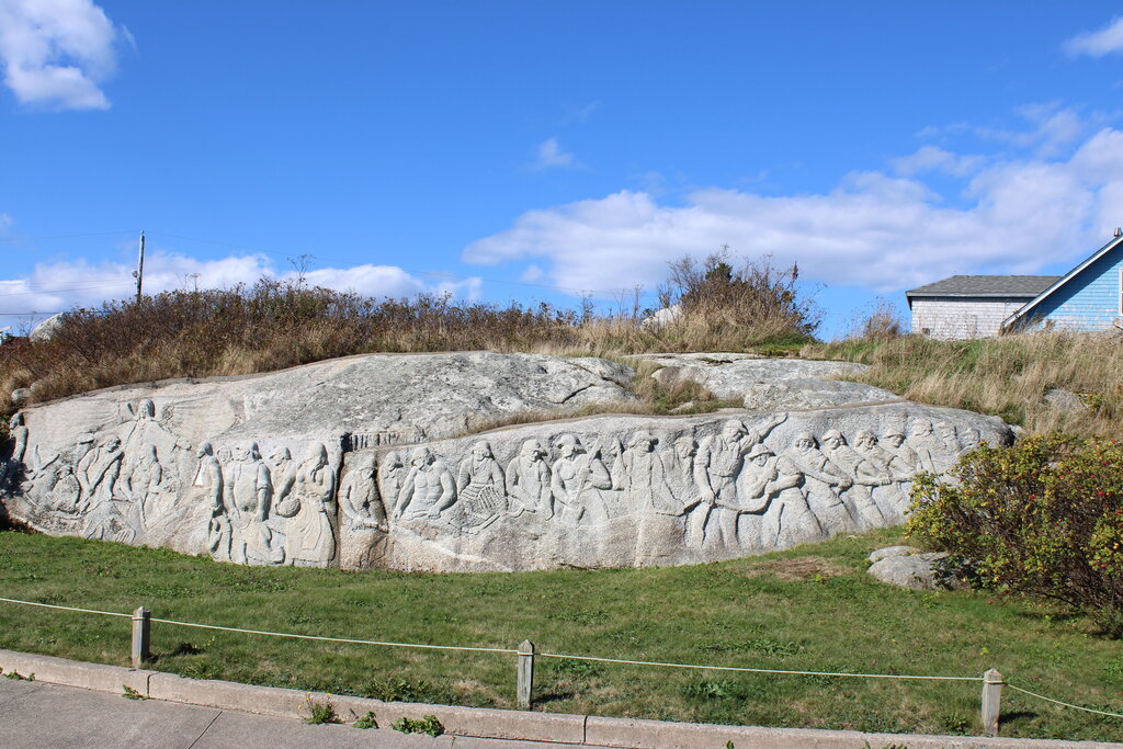 Large granite rock carved with detailed figures and scenes from coastal village life beside a grassy roadside. The carving stretches across the length of the rock under a blue sky.