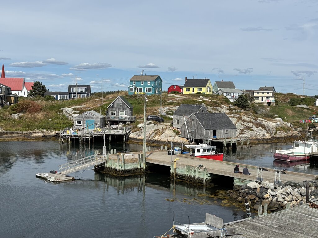 Colourful houses and weathered fishing sheds sit along a quiet harbour with calm water and small docks. A red fishing boat is moored near the shoreline beneath a cloudy sky.