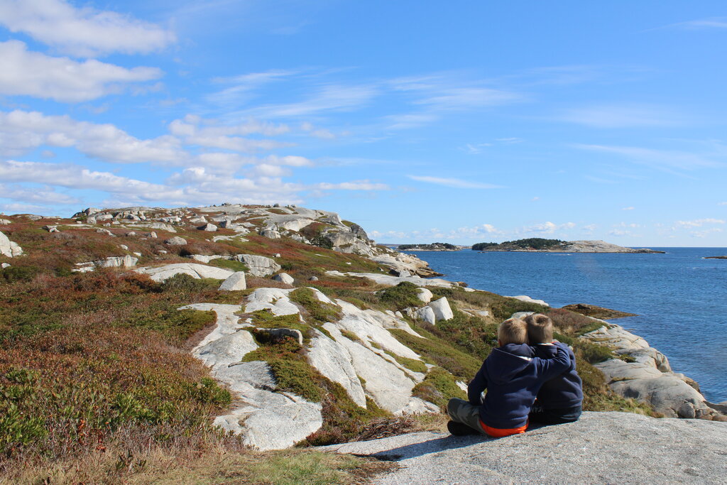 Two children sitting together on a rocky coastal cliff overlooking calm blue ocean water and small islands under a bright sky with scattered clouds. White granite rocks and low shrubs stretch across the shoreline around them.