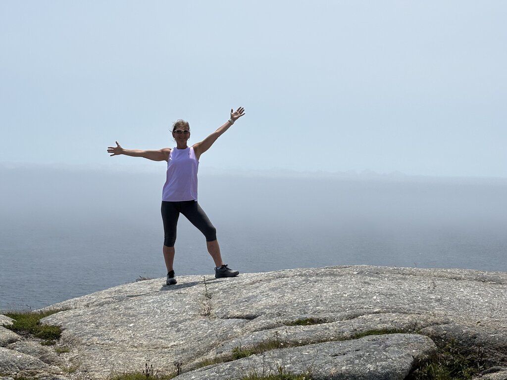 Woman standing with arms outstretched on a large rocky cliff overlooking the ocean in Nova Scotia. Hazy blue sky and calm water stretch across the background on a bright summer day.