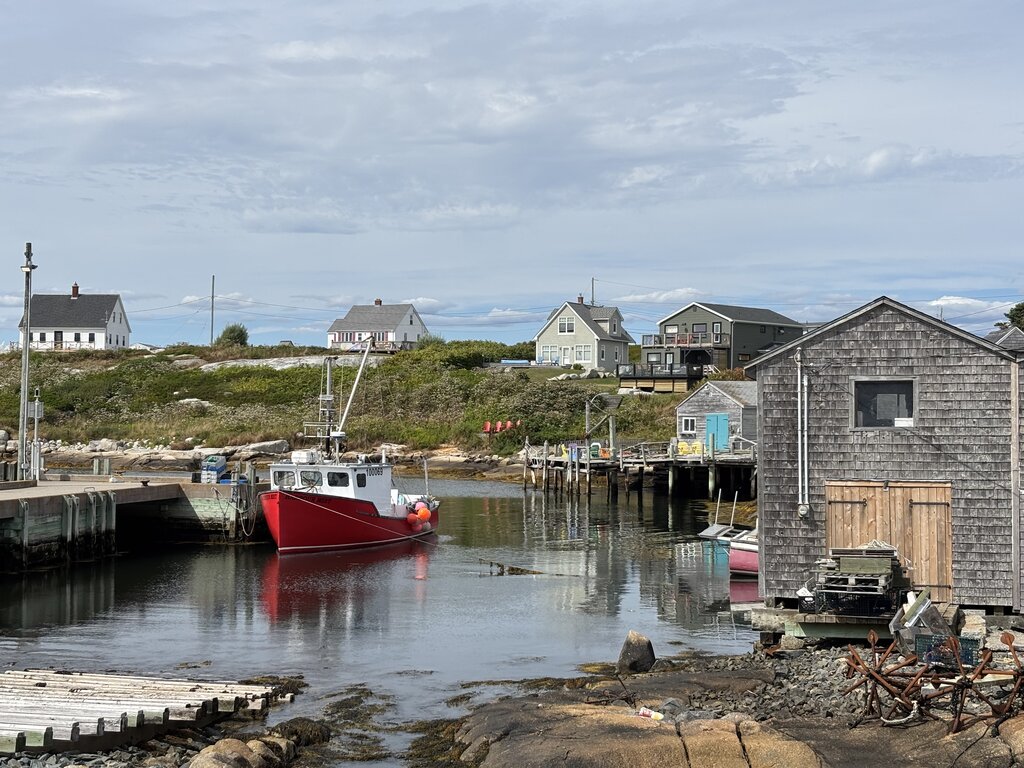 Bright red fishing boat floating in a narrow harbour in Peggy's Cove Nova Scotia surrounded by grey wooden sheds and coastal homes on rocky land. The still water reflects the buildings and boats along the shoreline.
