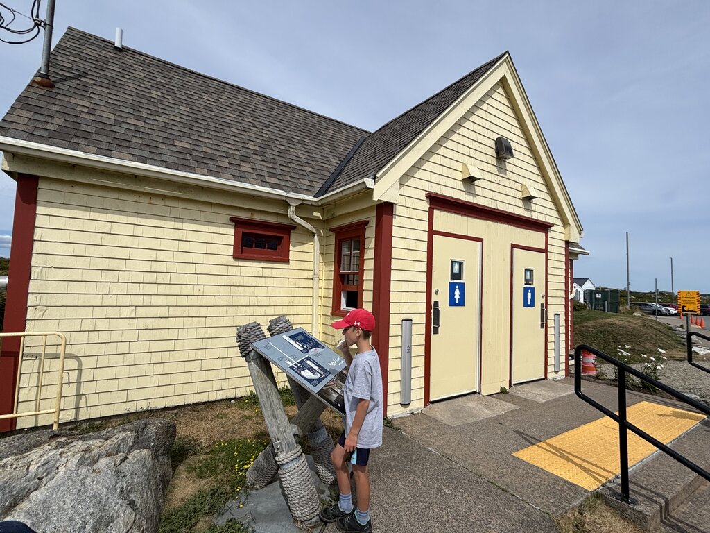 Child in a red hat reading an outdoor information panel beside a pale yellow coastal building with restroom signs on the doors. A rocky shoreline and walking paths extend into the background.