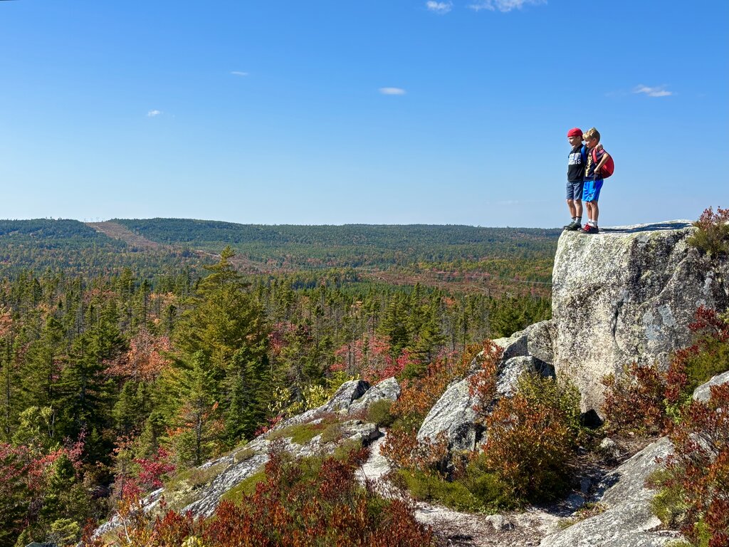 Two children stand on a large granite outcrop overlooking a wide forest beginning to turn red and orange beneath a bright blue sky. The rocky viewpoint highlights the colourful scenery found on fall hikes in Nova Scotia.