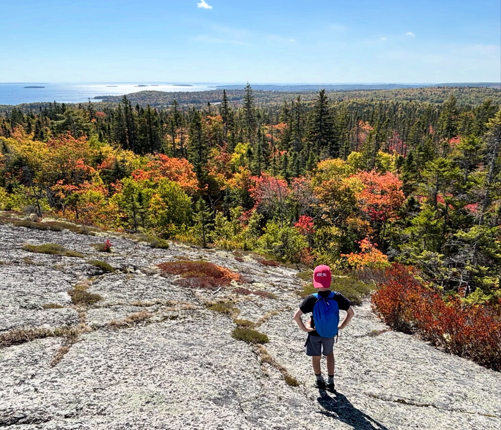 A child with a blue backpack stands on exposed granite above a forest filled with red orange yellow and evergreen trees. Water and small islands appear in the distance beyond the autumn landscape.